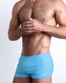 Side view of a masculine model wearing men’s swimsuit compression shorts in a solid light sky blue color made with Italian-made Vita By Carvico Econyl Nylon featuring a side pocket with official logo of BANG! Brand.