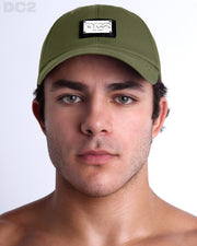 A man wearing the MAGNUM GREEN - Chillax Cap, a stylish deep forest green baseball cap made from breathable fabric. The cap features a metallic silver logo plaque on the front and a curved brim for sun protection.
