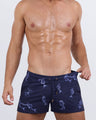 Frontal view of a male model wearing the MERMEN MASTERS Show Shorts by BANG! Miami. These navy blue swim shorts feature an intricate glowing mermaid and celestial star print, an elastic waistband with gold-tipped drawstrings, and a tailored short-leg fit.
