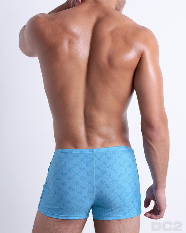 Back view of male model wearing the MONO BLUE beach sexy swimming bottoms for men in a blue color with a blue monogram logo, designed by DC2.