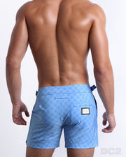Back view of male model wearing men’s MONO BLUE beach Tailored Shorts swimsuits in a light blue color with a darker blue monogram logo, complete with a back pocket, designed by DC2. 