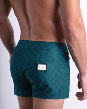 Back view of a male model wearing men’s Summer MONO TEAL Beach Shorts  in a teal color with a lighter blue monogram logo, complete with a back pocket, designed by DC2. made by DC2 a capsule brand by BANG! Clothes in Miami.