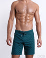 Male model wearing MONO TEAL beach Flex Boardshorts, premium swimwear in a dark teal green color with a darker teal color monogram logofor men. These high-quality swimwear bottoms by DC2, a men’s beachwear brand from Miami.