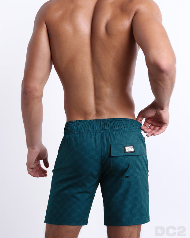 Back view of a male model wearing men’s MONO TEAL Flex Boardshorts swimsuits featuring the DC2 logo motif, these shorts were designed by DC2 in Miami.