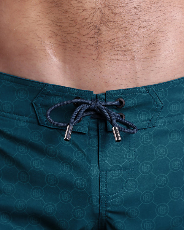 Close-up view of men’s summer long boardshorts by DC2 clothing brand, showing teal color cord with custom-branded golden cord ends, and matching custom eyelet trims in gold.
