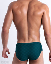 Back view of male model wearing the MONO TEAL beach Swim Briefs for men in a dark teal color with a blue monogram logo, designed by DC2.