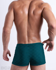 Back view of male model wearing the MONO TEAL beach sexy swimming bottoms for men in a dark teal color with a blue monogram logo, designed by DC2.