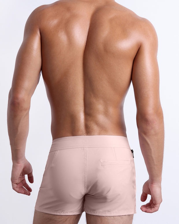 Male model wearing men’s NAKED PINK Beach Shorts swimsuit in a light rose quartz pink color, complete with a back pocket, designed by BANG! Clothes in Miami.