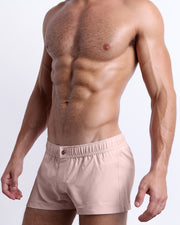 Side view of a masculine model wearing the men’s NAKED PINK shorter leg length swim shorts in a light pink color with the official logo of BANG! Brand. 