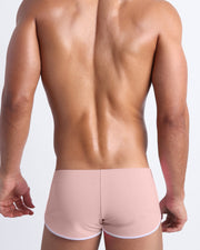 Back view of a male model wearing NAKED PINK men’s swim shorts in a light rose quartz pink color by the Bang! Clothes brand of men's beachwear.