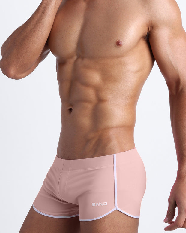 Side view of a masculine model wearing men’s swimwear in NAKED PINK a in a solid light pink color with official logo of BANG! Brand in white.