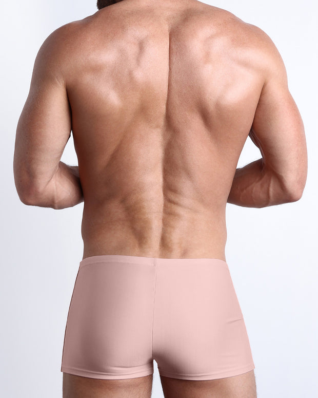 Back view of a male model wearing the NAKED PINK men’s swim trunks in a solid light pink color by the Bang! Clothes brand of men&