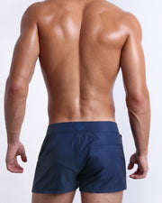 Male model wearing men’s NAVY BOOMER Beach Shorts swimsuit in a dark blue color, complete with a back pocket, designed by BANG! Clothes in Miami.