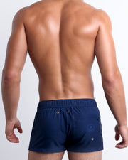 Back view of a male model wearing NAVY BOOMER men’s swim mini shorts with a back zipper pocket in a navy blue color by the Bang! Clothes brand of men's beachwear.