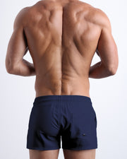Back view of male model wearing the NAVY BOOMER beach trunks in a dark blue color for men by BANG! Miami.
