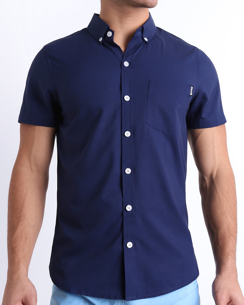 Front view of a sexy male model wearing NAVY BOOMER men’s short-sleeve stretch shirt in a dark denim blue color for men. This premium quality button-up top is by BANG! Clothes, a men’s beachwear brand from Miami.