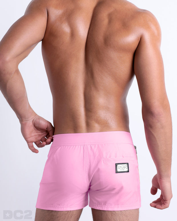 Back view of a male model wearing men’s Summer PADAM PINK Beach Shorts in a solid light pink color with pale yellow and aqua-blue side stripes, complete the back pockets, made by DC2 a capsule brand by BANG! Clothes in Miami.