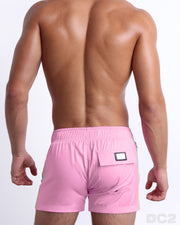 Back view of a male model wearing men’s PADAM PINK Flex Shorts swimsuits in a solid pink color with aqua and pastel yellow side stripes, complete the back pockets, made by DC2 a capsule brand by BANG! Clothes in Miami.