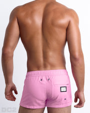 Back view of male model wearing men’s PADAM PINK beach Mini Shorts swimsuits in a solid pink color with side stripes in blue and pastel yellow colors, complete with a back pocket, designed by DC2 a capsule brand by BANG! Clothes based in Miami.