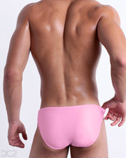 Back view of male model wearing the PADAM PINK beach mini-briefs for men in a solid pink color with side blue and yellow stripes, designed by DC2.