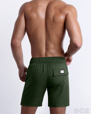 Back view of male model wearing men’s PALM GREEN beach Flex Boardshorts swimming shorts. In a solid moss green color, complete with a back pocket, designed by DC2 a capsule brand by BANG! Clothes based in Miami.