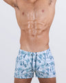 Frontal view of a male model wearing the Party Animals Show Shorts for men by BANG! Miami. These premium short-length beach shorts feature a soft white base with an intricate blue toile-inspired print depicting exotic animals and tropical scenery. Designed with a tailored fit, an adjustable drawstring waistband with metallic tips, and a modern athletic cut for a confident beachwear look.