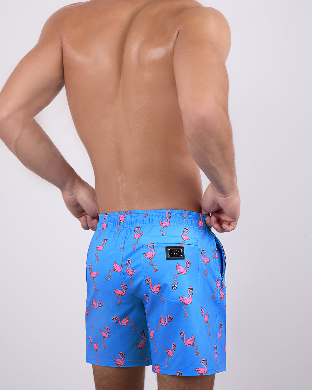 Back view of a male model wearing the PINK FLAMINGOS Resort Shorts by DC2 Miami. These resort-style swim shorts showcase a tailored, long-leg cut, a functional back pocket with a DC2 signature metal logo plaque, and a breathable, quick-dry fabric for ultimate comfort.