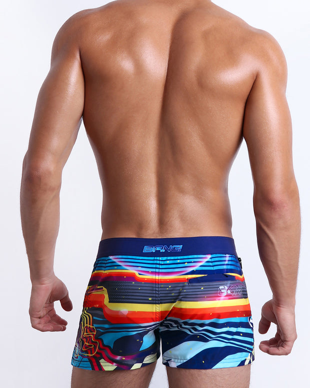 Back view of male model wearing men’s POOL POSITION Beach Shorts swimsuit, a racing-inspired design with a burst of colors including blue, red, yellow, and orange is designed by BANG! Clothes in Miami.