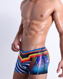 Side view of muscular male model wearing POOL POSITION Summer swimming trunks swimsuit for the beach in a racing-inspired design with a burst of colors including blue, red, yellow, and orange is designed by BANG! Clothes in Miami.