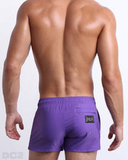 Male model wearing men’s PURPLE MACHINE Summer Poolside Shorts swimsuit in a solid purple color, complete with a back metallic zippered-pocket, designed by DC2 a capsule brand by BANG! Clothes based in Miami.