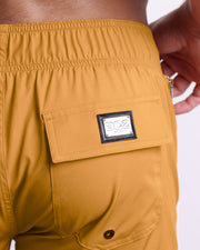 Close-up view of the RETRO MUSTARD men’s Flex Boardshorts back pocket, showing custom branded silver metal logo.