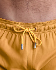 Close-up view of the RETRO MUSTARD men’s summer shorts, showing dark yellow cord with custom branded silver cord ends, and matching custom eyelet trims in silver.
