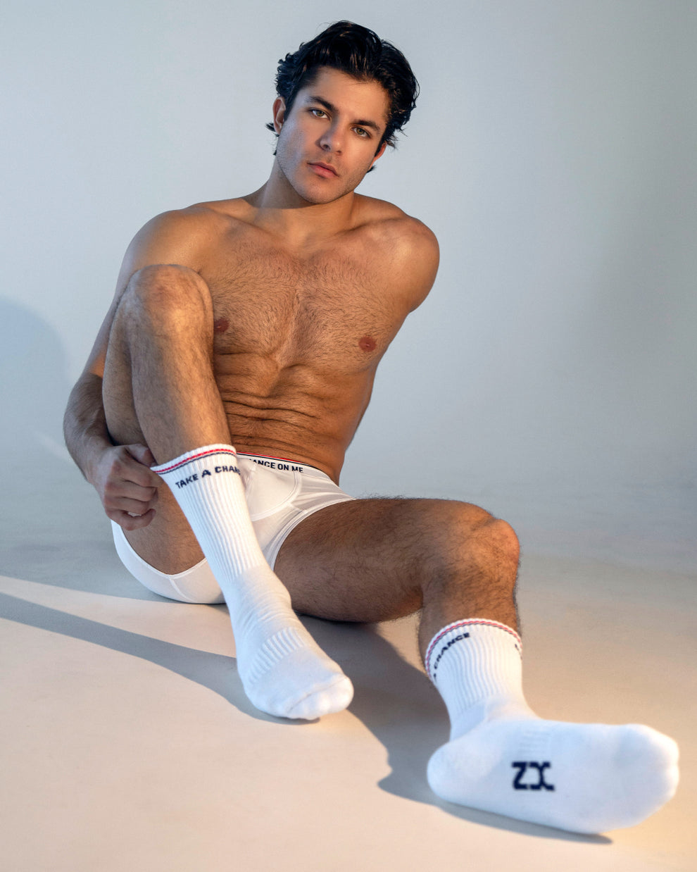 DC2 TAKE A CHANCE ON ME Boxer Brief — male model seated in studio wearing white men’s modal boxer briefs with logo waistband, paired with white crew socks, showing relaxed lifestyle look.