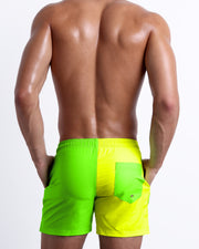 Back view of a male model wearing men’s SINGLE BILINGUAL (NEON YELLOW/GREEN) beach Resort Shorts swimsuits in a bright fluorescent yellow and green color block design made by BANG! Clothes in Miami.
