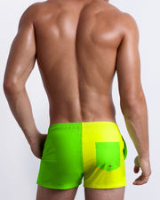 Back view of a male model wearing men’s SINGLE BILINGUAL (NEON YELLOW/GREEN) beach Show Shorts swimsuits in a bright fluorescent yellow and green color block design made by BANG! Clothes in Miami.
