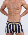 Frontal view of a masculine model wearing the SUN CHASING STRIPES Mini Shorts for men by DC2 Miami, a premium swimwear brand. These ultra-short beach shorts feature a bold navy blue base with striking yellow and white stripes, an elastic waistband with an adjustable internal drawstring, and a sleek fitted cut, making them perfect for stylish summer looks, beach days, and poolside lounging.