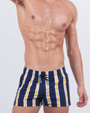 Model wearing SUN CHASING STRIPES Poolside swimming shorts for men, featuring bold navy, white, and yellow vertical stripes. Statement resortwear with a quad-hugging silhouette and metallic drawcord tips for a sporty-luxe finish.