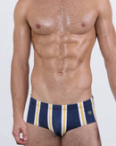 Model wearing SUN CHASING STRIPES men’s swim sunga featuring a bold navy base with vertical golden-yellow and white stripes. This Brazilian-style swimsuit delivers a sleek fit with confident energy and vibrant resort style.