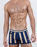 Model wearing SUN CHASING STRIPES swim trunks in a bold navy base with vertical white and golden yellow stripes. Men's designer swimwear with a skin-hugging square-cut fit and a wide contrast waistband for modern resort style.