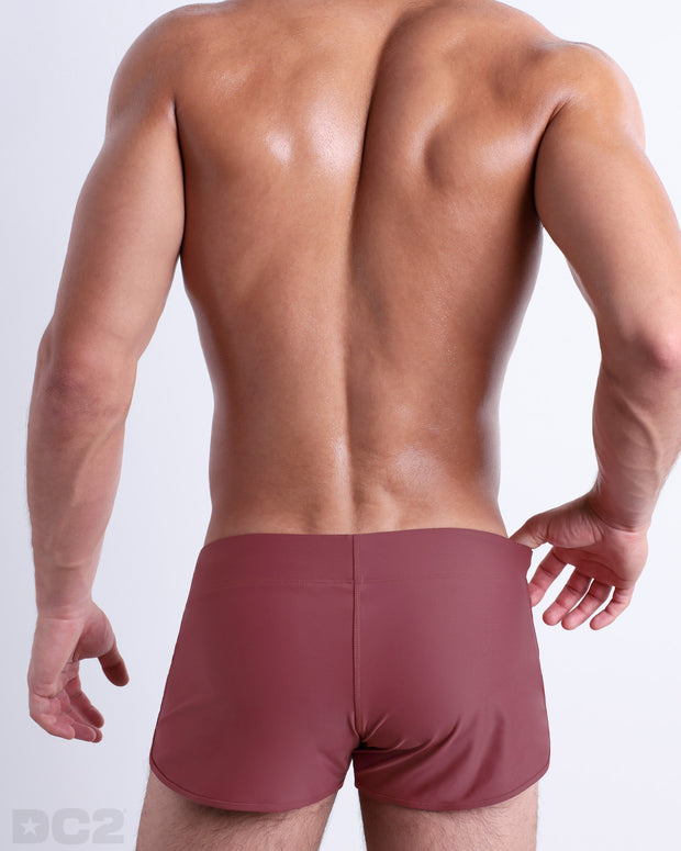 Back view of male model wearing the SUN-KISSED RED beach Swim Shorts for men by BANG! Miami in a solid blush color.