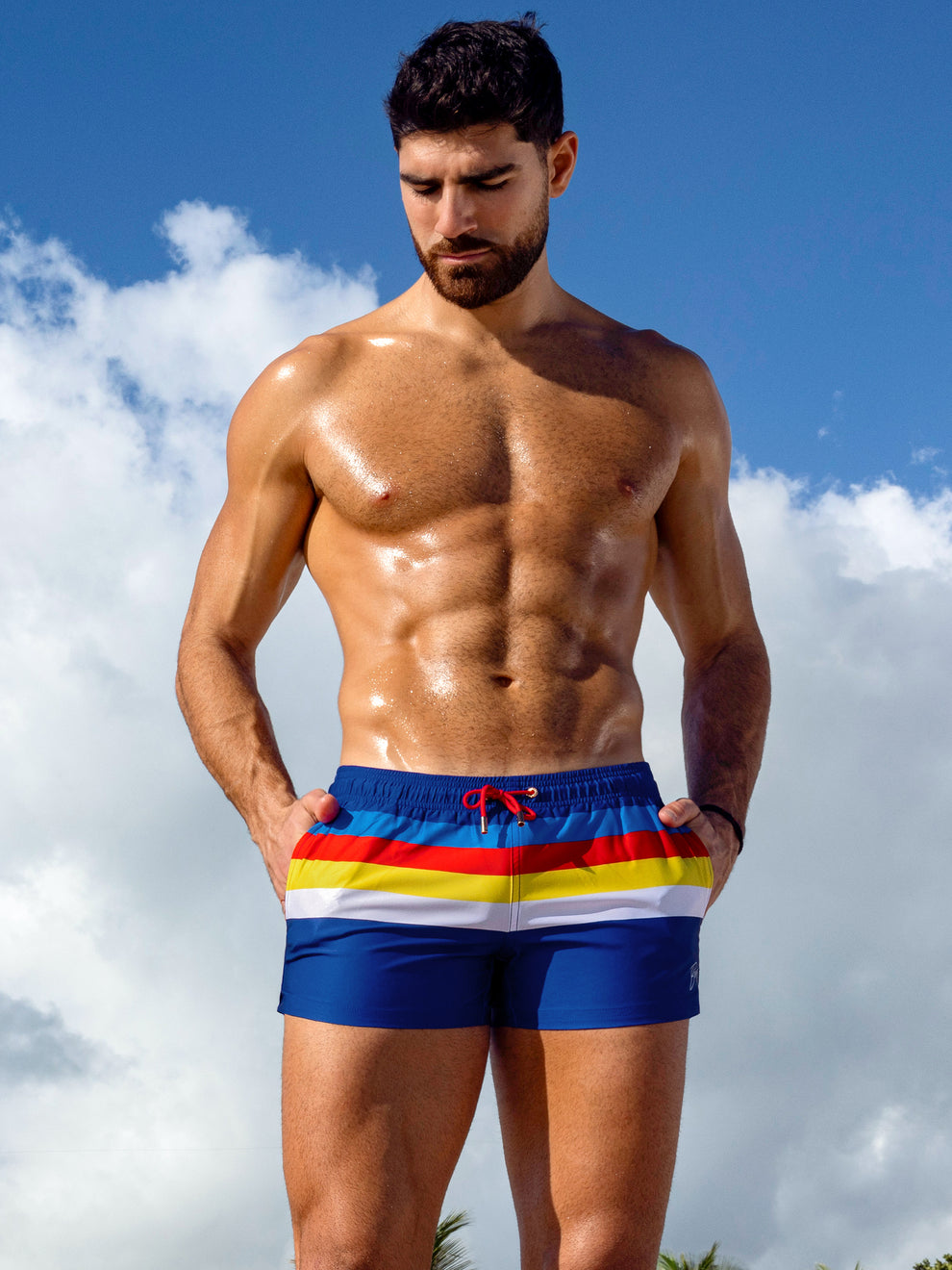 Model wearing Stripe'a'Pose Show Shorts with bold blue, white, yellow and red horizontal stripes, featuring a red drawstring and retro-inspired swim style.