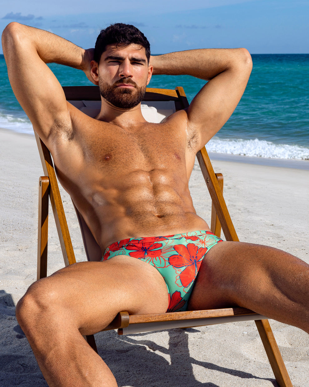 Model reclining on a beach chair wearing aqua swim briefs with bold red floral print, set against a sunny ocean backdrop.