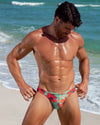 Male model wearing the Surf Flowers (Aqua/Red) men’s Swim Mini Brief in a vibrant floral print on the beach.