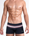 Model wearing The Ilya Swim Trunks in black with red, white, and gray stripe detailing, featuring a sleek square-cut silhouette and premium men's designer swimwear.