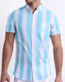 Male model wearing THE KEN (MYKONOS EDITION) men’s sleeveless stretch shirt, in white and light blue-colored stripes inspired by the styles seen worn by Ryan Gosling as Ken, in the Barbie movie. Designed by BANG! Clothes, a men’s beachwear brand from Miami.