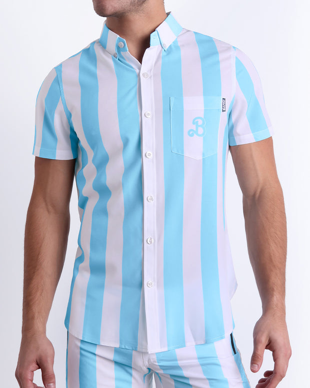 Male model wearing THE KEN (MYKONOS EDITION) men’s sleeveless stretch shirt, in white and light blue-colored stripes inspired by the styles seen worn by Ryan Gosling as Ken, in the Barbie movie. Designed by BANG! Clothes, a men’s beachwear brand from Miami.
