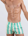Frontal view of a male model wearing THE KEN (TULUM EDITION) Show Shorts for men by BANG! Miami. These stylish above-the-knee beach shorts feature bold green and cream vertical stripes, an adjustable drawstring waistband with gold-tone details, and a contoured fit designed for comfort and movement.