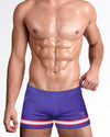 Model wearing The Shane Swim Trunks in purple with red and white stripe detailing, featuring a modern square-cut silhouette and premium men's designer swimwear.
