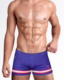 Model wearing The Shane Swim Trunks in purple with red and white stripe detailing, featuring a modern square-cut silhouette and premium men's designer swimwear.