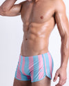 Side view of a male model wearing men’s THE KEN (IBIZA EDITION) sexy men’s swimming shorts featuring pastel pink and light blue stripes is designed by BANG! Clothes in Miami.